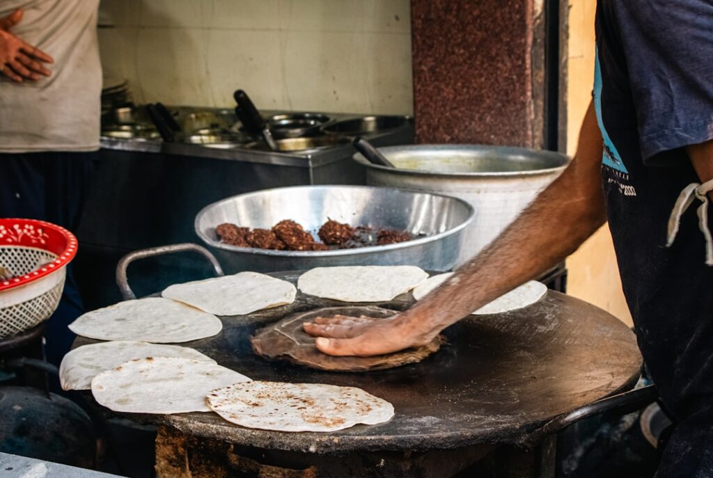 A street vendor in Mumbai preparing Indian flatbread on a large, blackened tawa or griddle. The vendors arms, dusted with flour, handle a partially cooked, round bread, emphasizing the texture of the dough against the rough iron surface. The ambient light highlights the simple, rustic process of cooking, making it ideal for content related to street food, culinary traditions, travel in India, or authentic cooking blogs and articles, conveying a sense of immediacy and cultural authenticity.