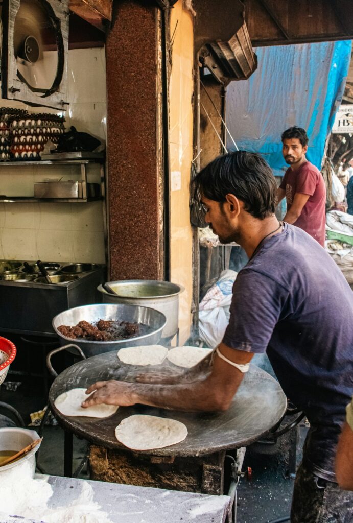 A street vendor in Mumbai preparing Indian flatbread on a large, blackened tawa or griddle. The vendors arms, dusted with flour, handle a partially cooked, round bread, emphasizing the texture of the dough against the rough iron surface. The ambient light highlights the simple, rustic process of cooking, making it ideal for content related to street food, culinary traditions, travel in India, or authentic cooking blogs and articles, conveying a sense of immediacy and cultural authenticity.