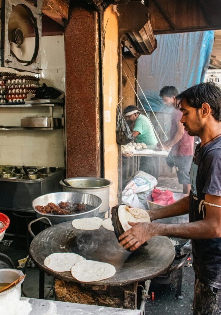 A street vendor in Mumbai preparing Indian flatbread on a large, blackened tawa or griddle. The vendors arms, dusted with flour, handle a partially cooked, round bread, emphasizing the texture of the dough against the rough iron surface. The ambient light highlights the simple, rustic process of cooking, making it ideal for content related to street food, culinary traditions, travel in India, or authentic cooking blogs and articles, conveying a sense of immediacy and cultural authenticity.